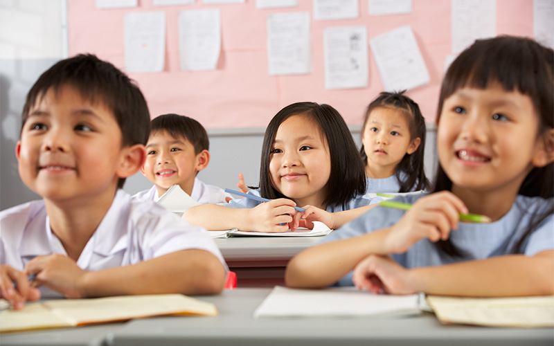 A group of young school children sitting at desks in a classroom, looking forward attentively.