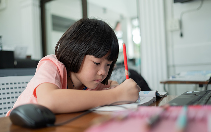 A young student with short dark hair in a pink shirt focuses intently on writing in a notebook next to a computer mouse.