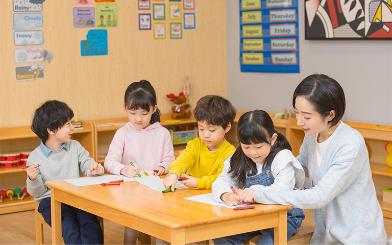 A teacher helps four young children as they draw with crayons at a wooden table in a brightly decorated classroom.
