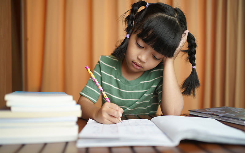 A young girl with pigtails in a green striped shirt rests her head on her hand while writing in a workbook.