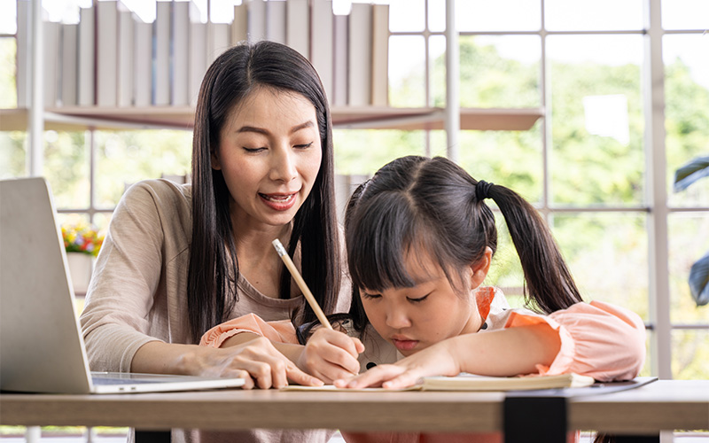 A woman sitting at a desk and guiding a young girl with pigtails as she writes in a notebook.