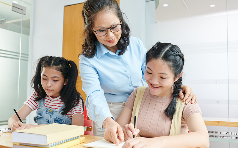 An older female educator leaning over to provide guidance to two young students working on their lessons.