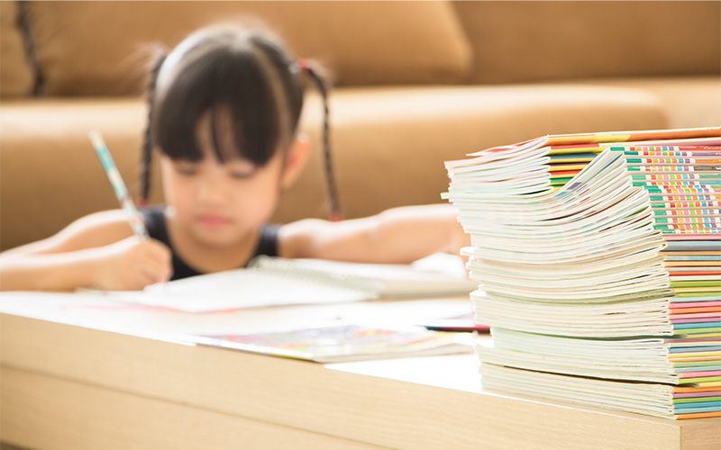 A young student with pigtails writing in a notebook, viewed behind a large, tall stack of colorful books.