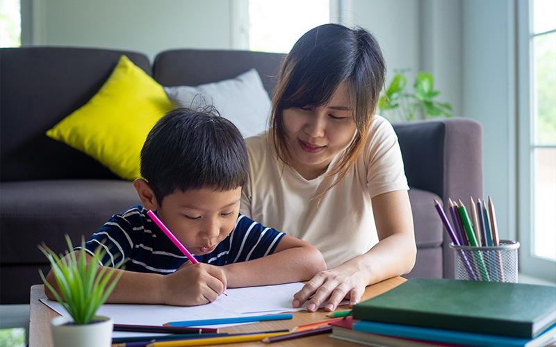 Adult guiding a young child as he writes or draws at a table, with colored pencils, books, and a cozy living room in the background