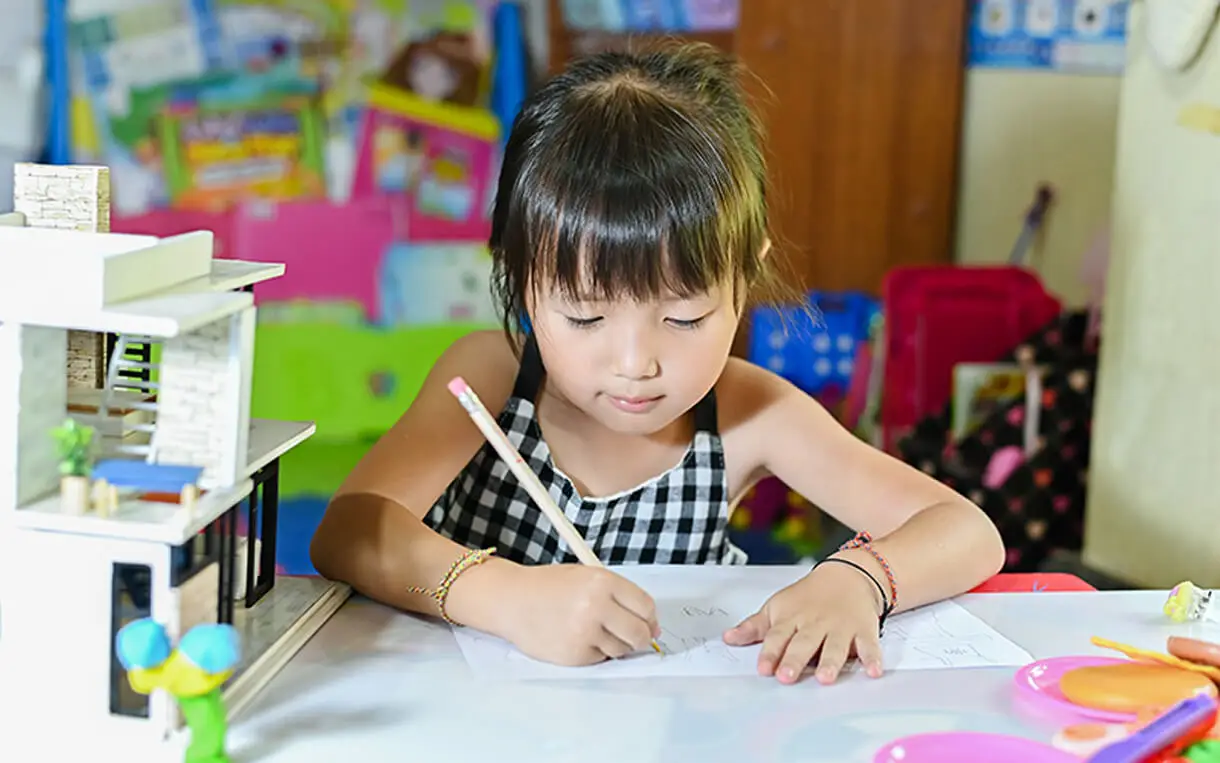 Young child drawing on paper at a desk with a model house and art supplies nearby