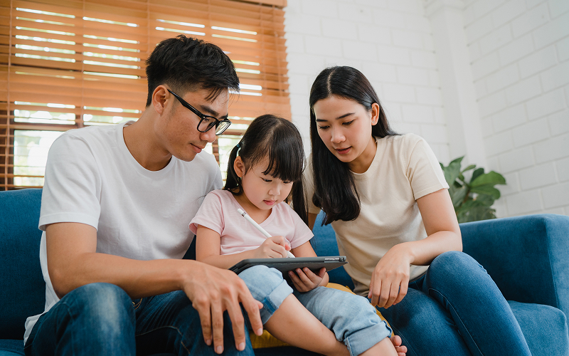 Parents watching while child uses tablet on blue sofa.