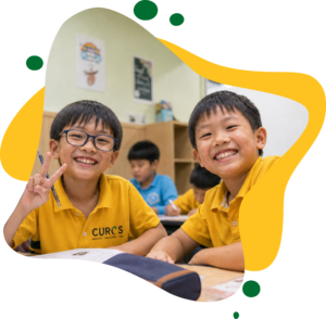 Two smiling schoolboys in CUROS uniforms pose at a classroom desk; one flashes a peace sign, with classmates working behind them.